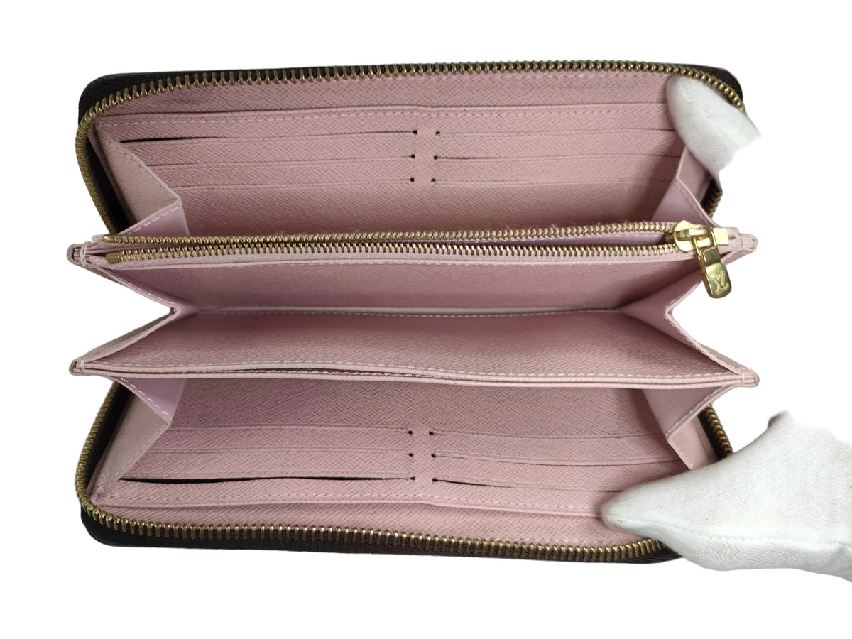 An open pink leather zip-around wallet with multiple card slots and compartments, held by a white gloved hand. The wallet features a dark brown monogram pattern along the outer edges and gold-tone zippers.