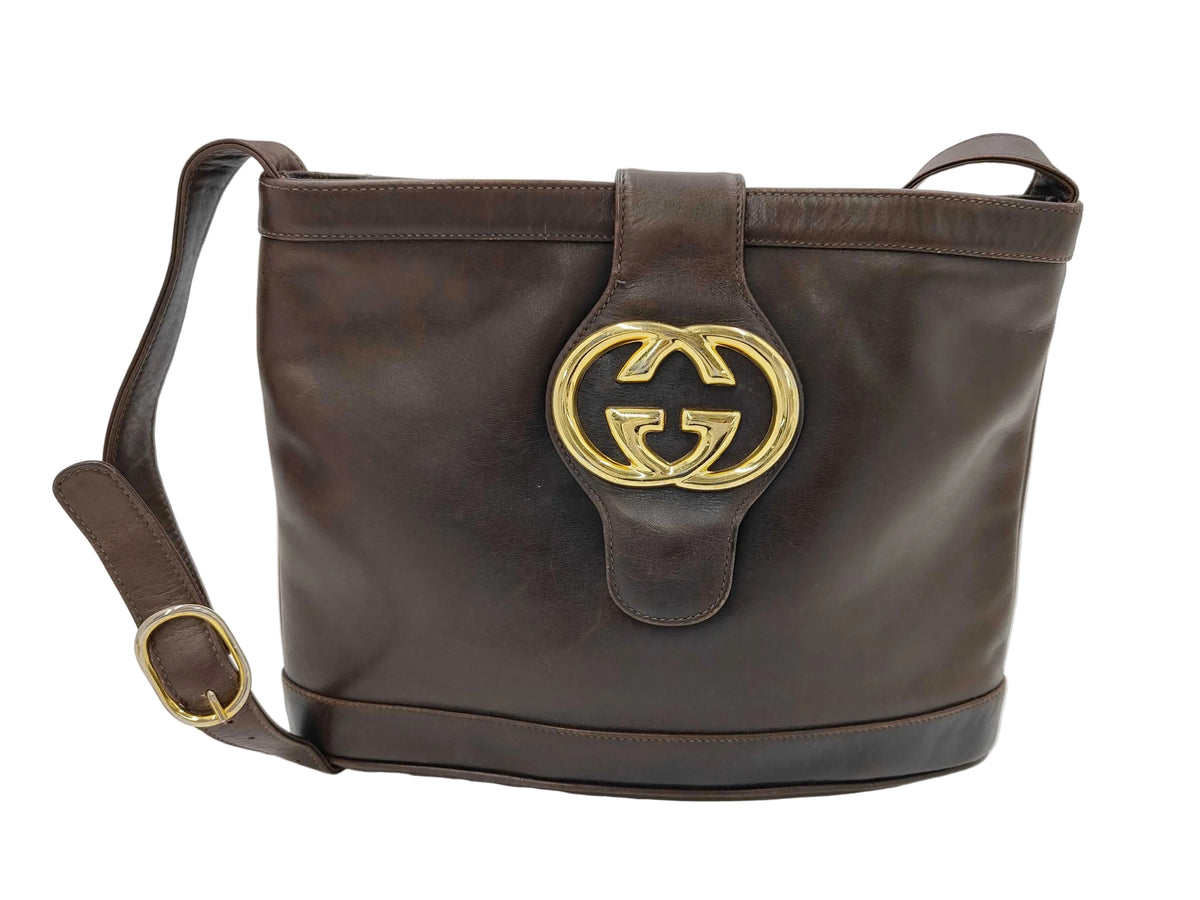 A dark brown leather shoulder bag with a large gold-tone interlocking G logo on the front, indicating the brand Gucci. The bag features a top handle, a detachable shoulder strap with a gold buckle, and a structured silhouette, photographed against a white studio background.