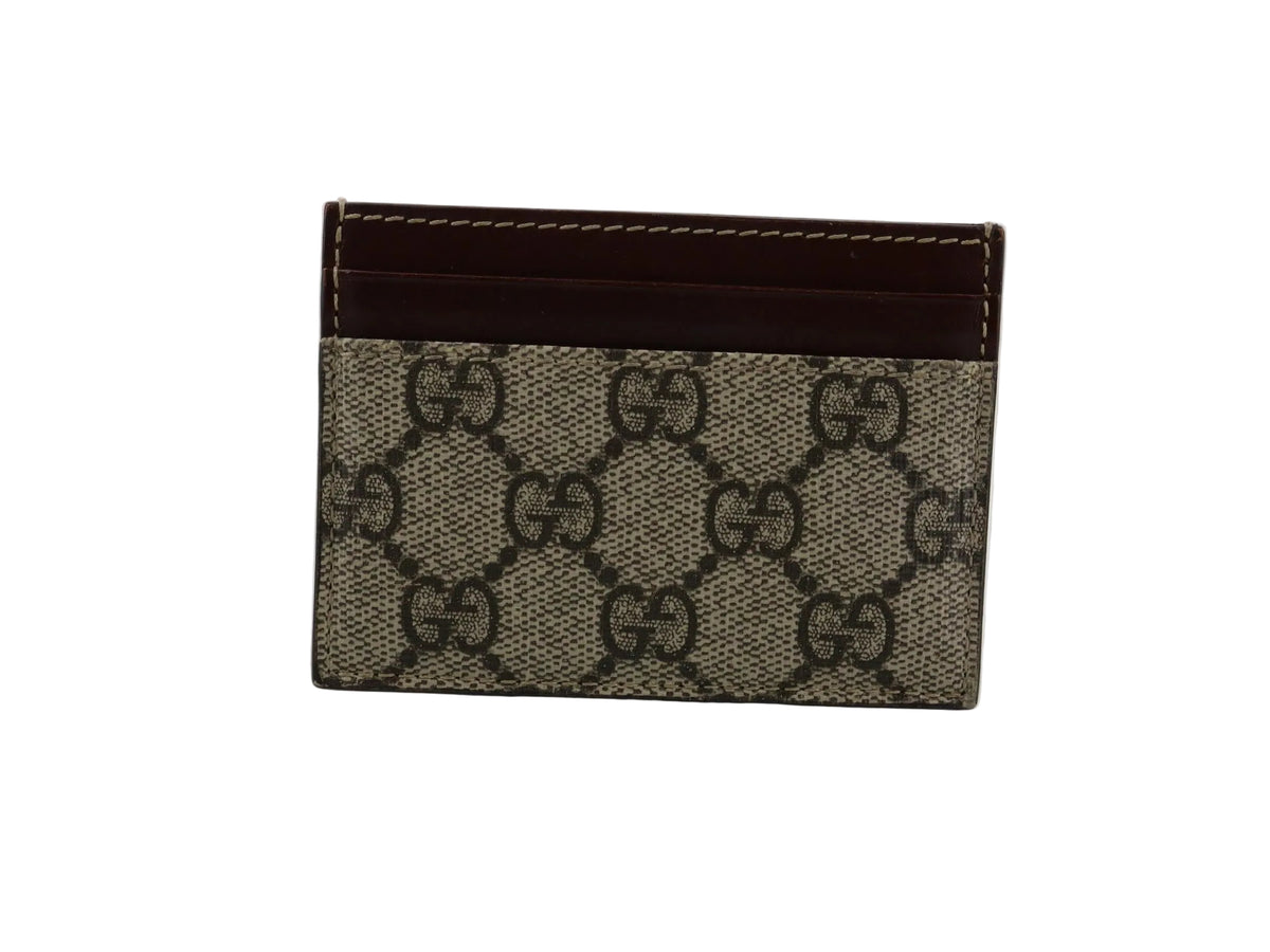 A small Gucci card holder wallet in beige and brown GG Supreme canvas with a dark brown leather top band, shown standing upright on a light reflective surface.